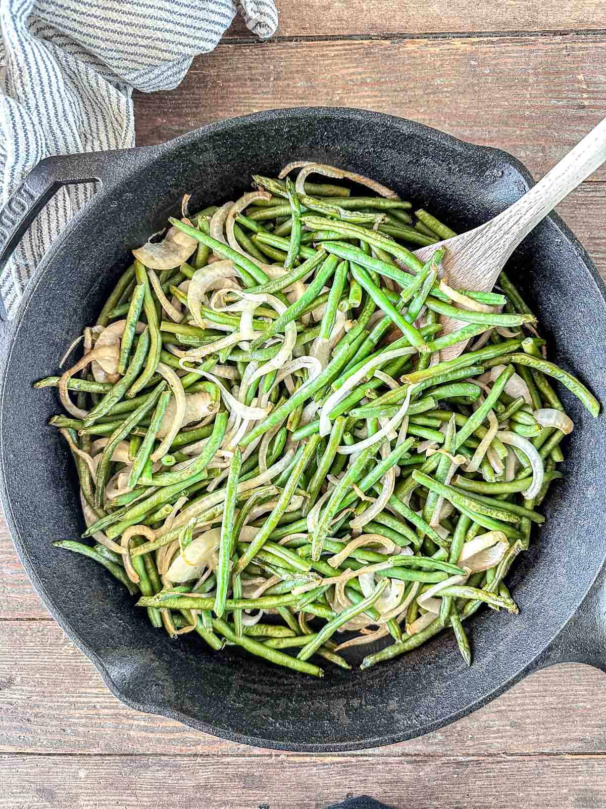 A cast iron skillet filled with sautéed green beans and sliced onions, mixed with a wooden spoon, sits on a wooden surface next to a striped cloth.
