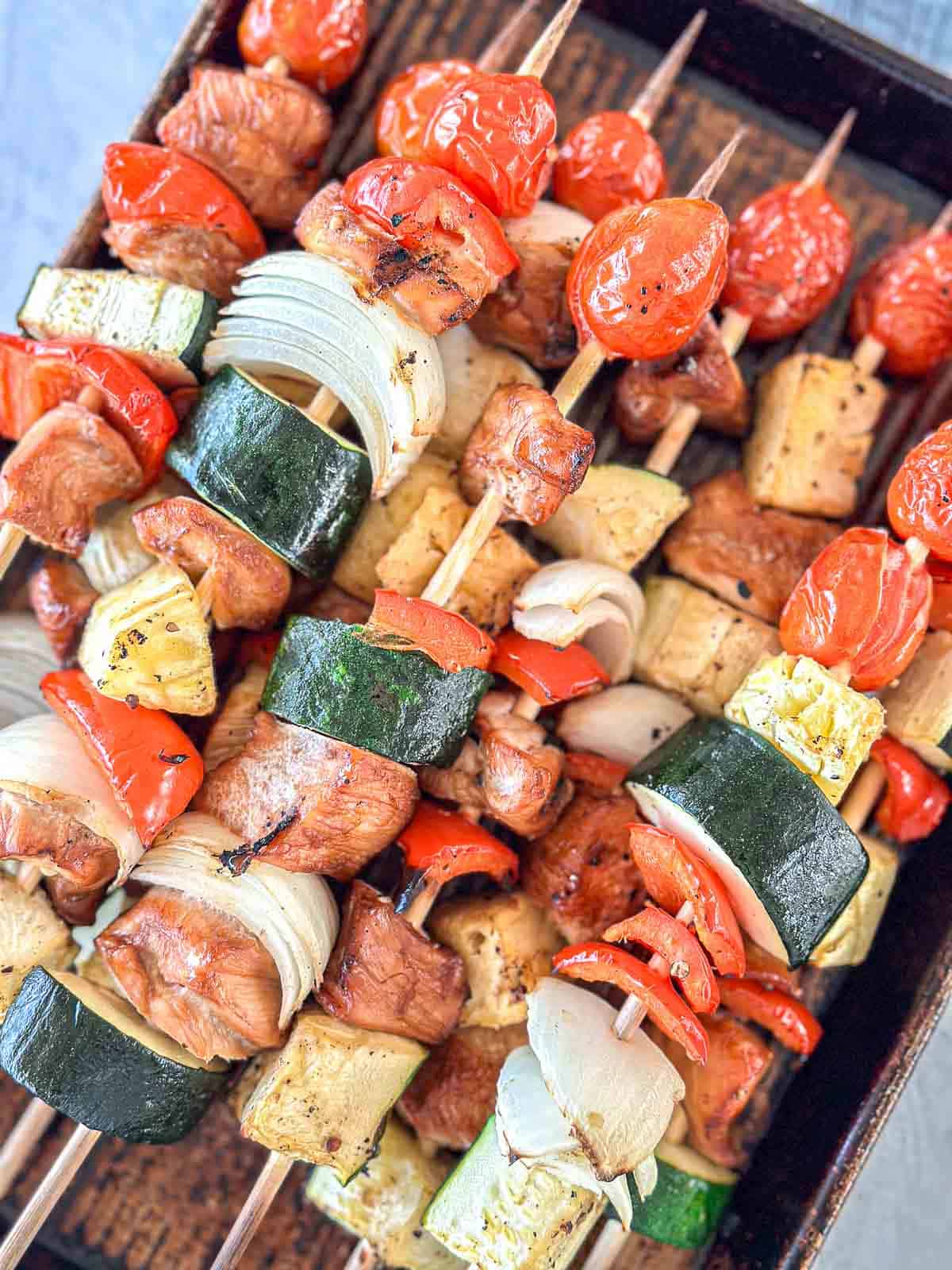 Wooden skewers with grilled chicken, cherry tomatoes, zucchini, onion, red bell pepper, and pineapple chunks are arranged on a baking tray. The vegetables and chicken are evenly spaced and appear freshly cooked.
