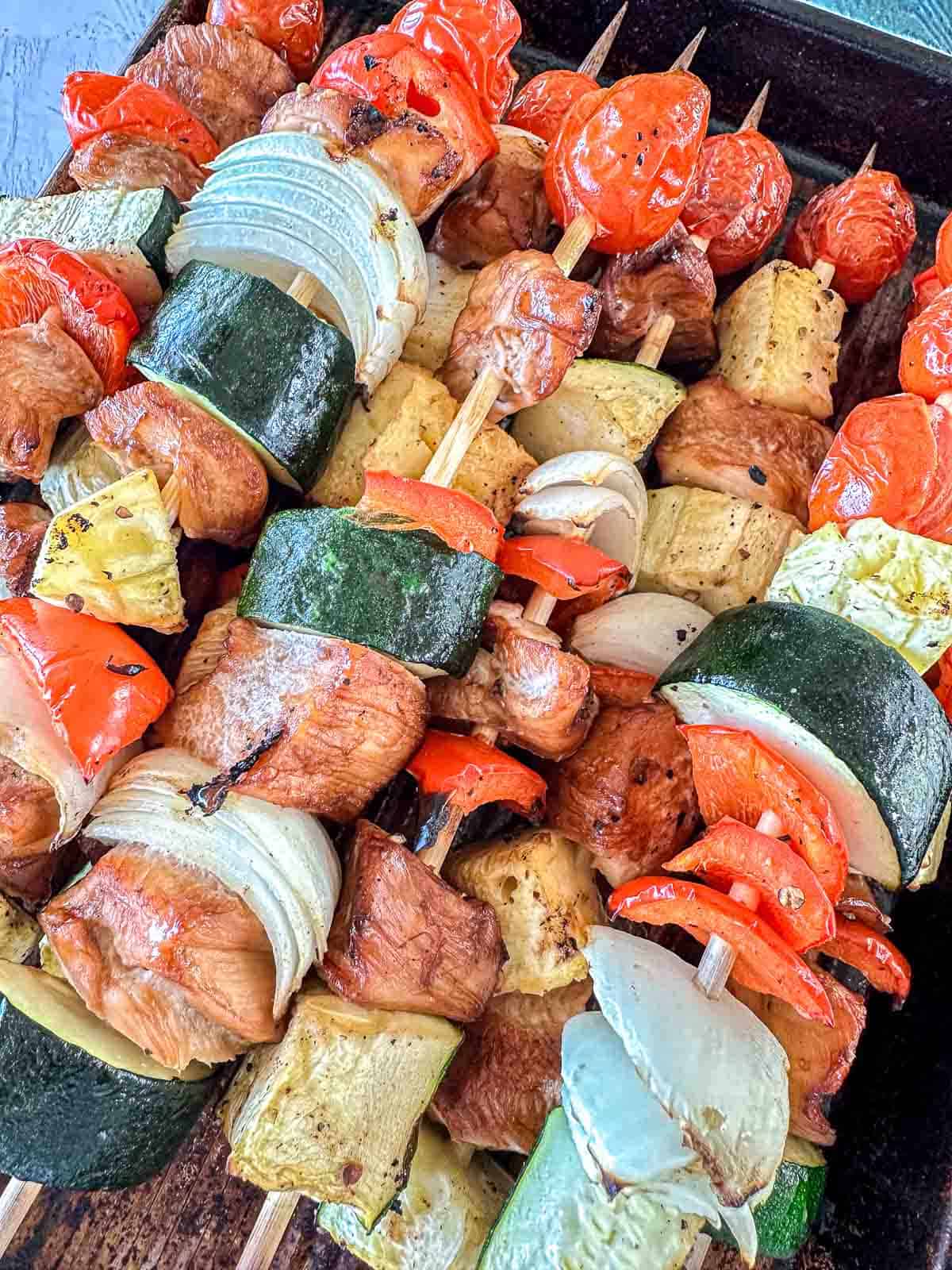A close-up of grilled skewers loaded with chunks of chicken, zucchini, onions, cherry tomatoes, red bell peppers, and yellow squash on a dark tray.
