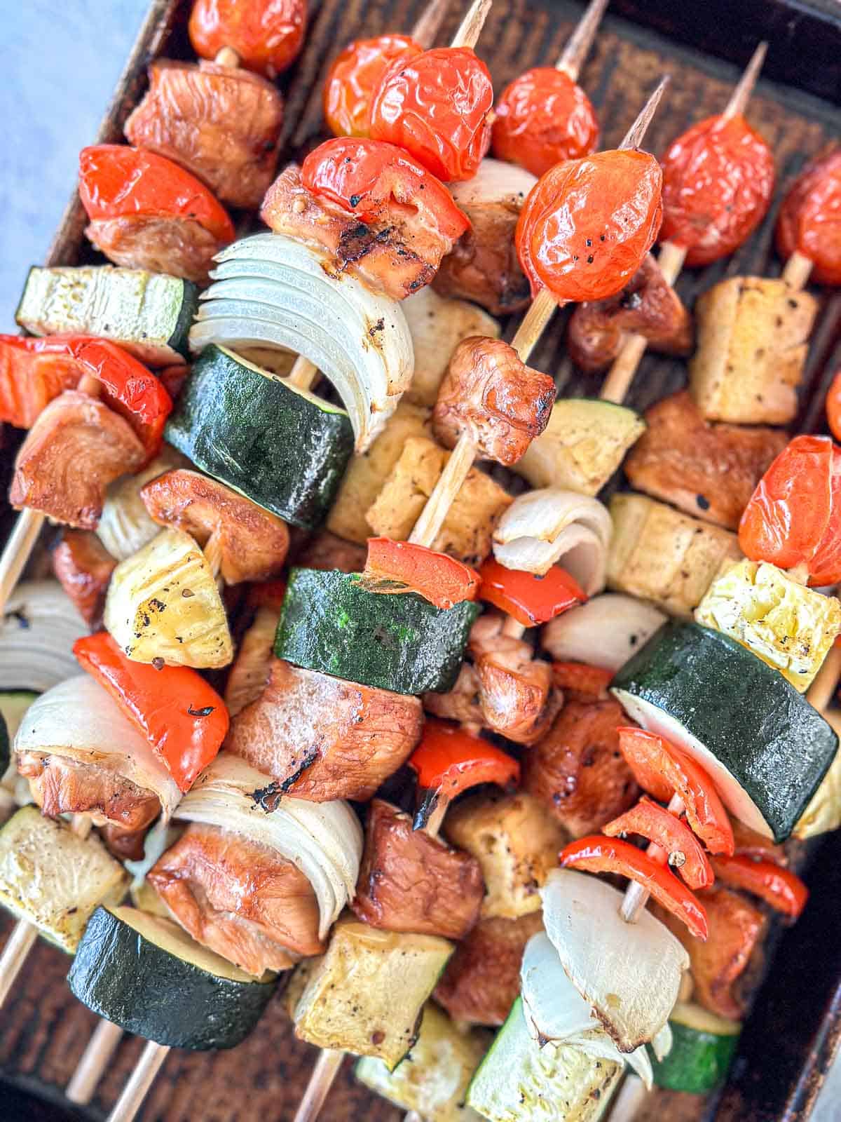 Skewers loaded with grilled chicken, zucchini, onion, red bell pepper, tomatoes, and pineapple slices, arranged closely on a tray. The vegetables and meat are lightly charred and colorful.