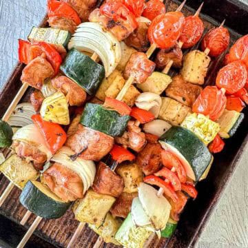 Chicken Skewers filled with grilled vegetables such as zucchini, onion, cherry tomatoes, red bell pepper, and chunks of chicken and pineapple, arranged on a dark baking tray.