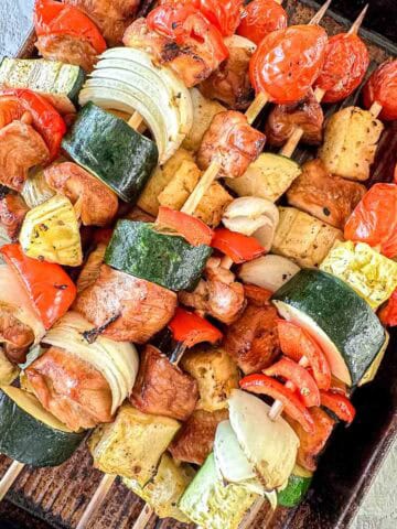 Chicken Skewers filled with grilled vegetables such as zucchini, onion, cherry tomatoes, red bell pepper, and chunks of chicken and pineapple, arranged on a dark baking tray.