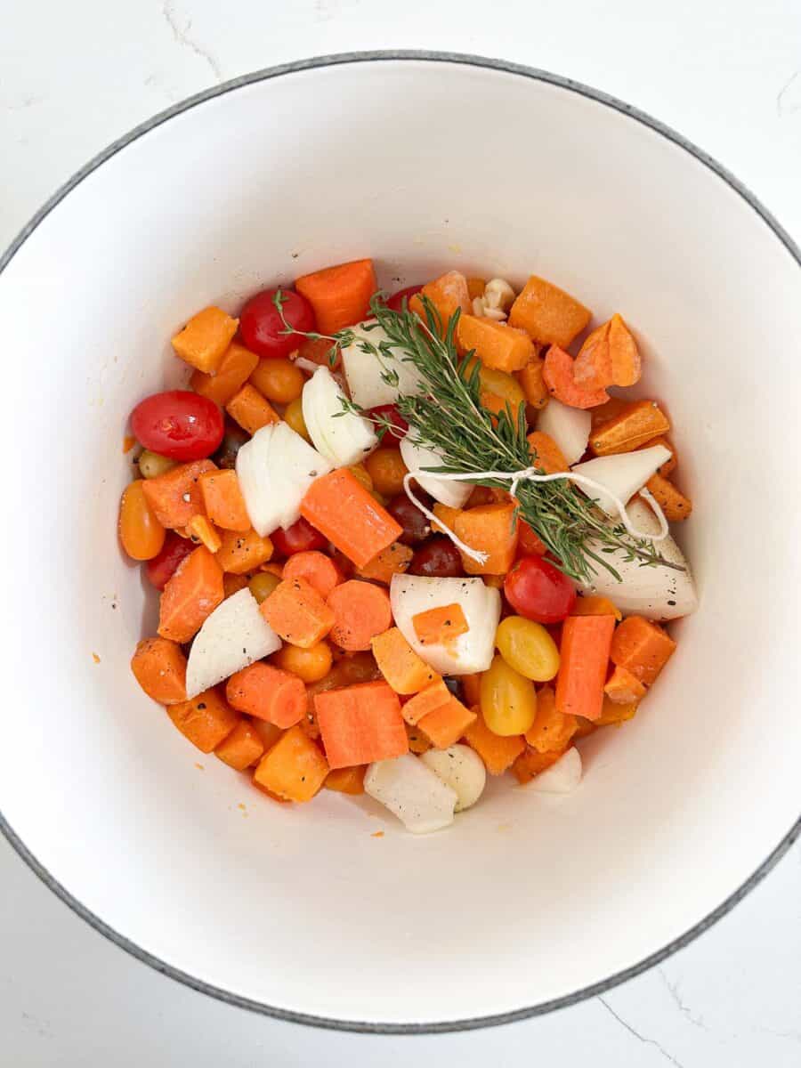A white Dutch oven filled with butternut squash, tomatoes, onion, garlic and herbs.