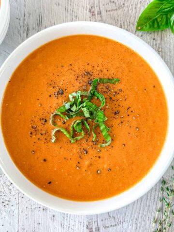 Butternut Squash And Tomato Soup in a white bowl topped with fresh herbs and black pepper.