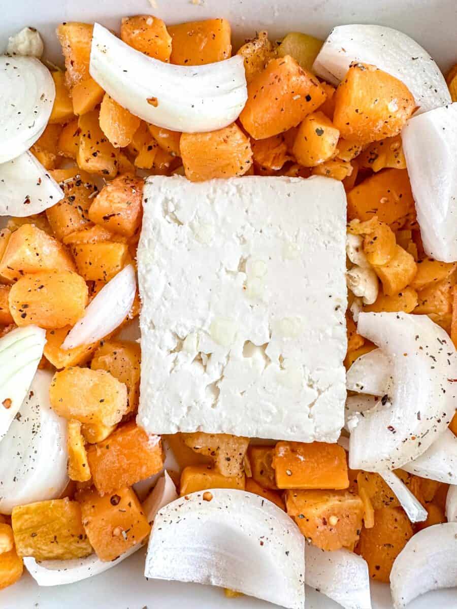 A block of feta cheese sits in the center of a baking dish surrounded by chopped butternut squash and onion pieces, all seasoned with pepper and spices.
