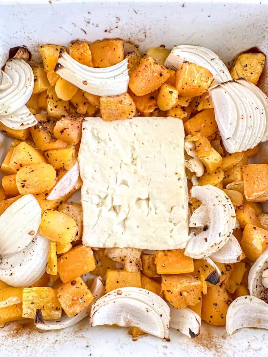 Cubed butternut squash and sliced onions are arranged in a baking dish around a large block of feta cheese, all seasoned with black pepper and roasted.