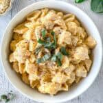 A white bowl filled with butternut squash feta pasta shells topped with fresh sage leaves and cracked black pepper, placed on a light-colored wooden surface.