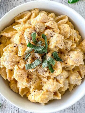 A white bowl filled with butternut squash feta pasta shells topped with fresh sage leaves and cracked black pepper, placed on a light-colored wooden surface.