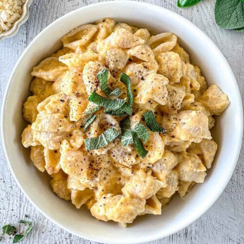 A white bowl filled with butternut squash feta pasta shells topped with fresh sage leaves and cracked black pepper, placed on a light-colored wooden surface.