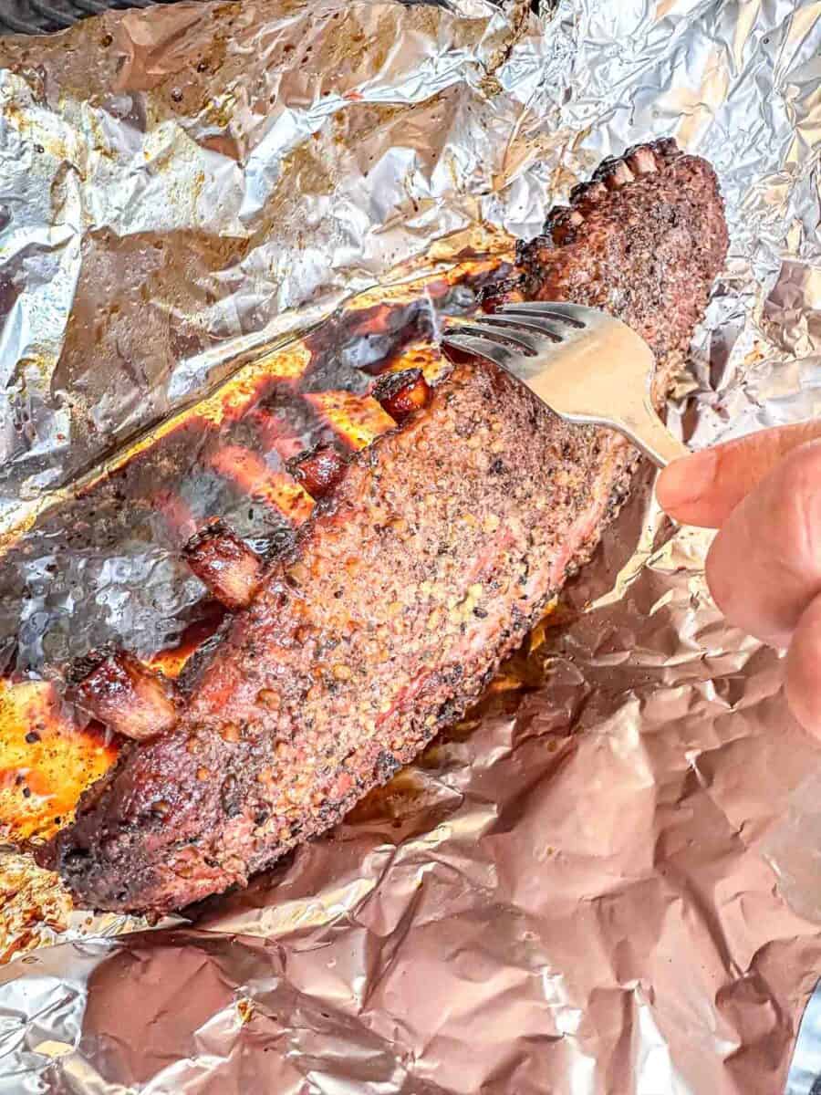 Baby back ribs in foil. There is a fork showing how far the meat has pulled back from the bones.