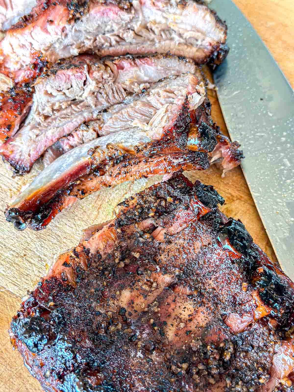 Close-up of sliced, grilled beef ribs on a wooden cutting board, showing a peppery, charred crust and juicy, tender meat. A large knife is positioned beside the slices.