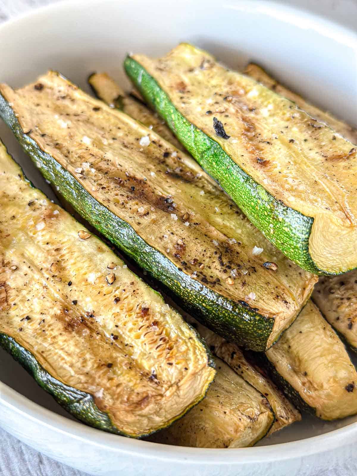 A white bowl filled with roasted zucchini slices seasoned with black pepper and salt. The zucchini pieces are golden brown and arranged neatly in the bowl.