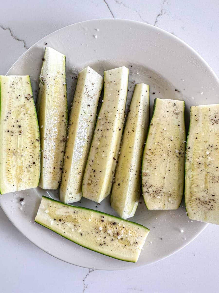 A plate with sliced zucchini on it. They are covered with oil, salt and pepper.