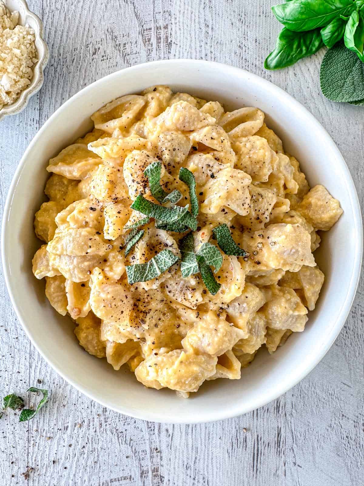 A bowl of butternut squash feta pasta shells topped with chopped fresh herbs and cracked black pepper, placed on a light wooden surface with grated cheese and fresh basil on the side.