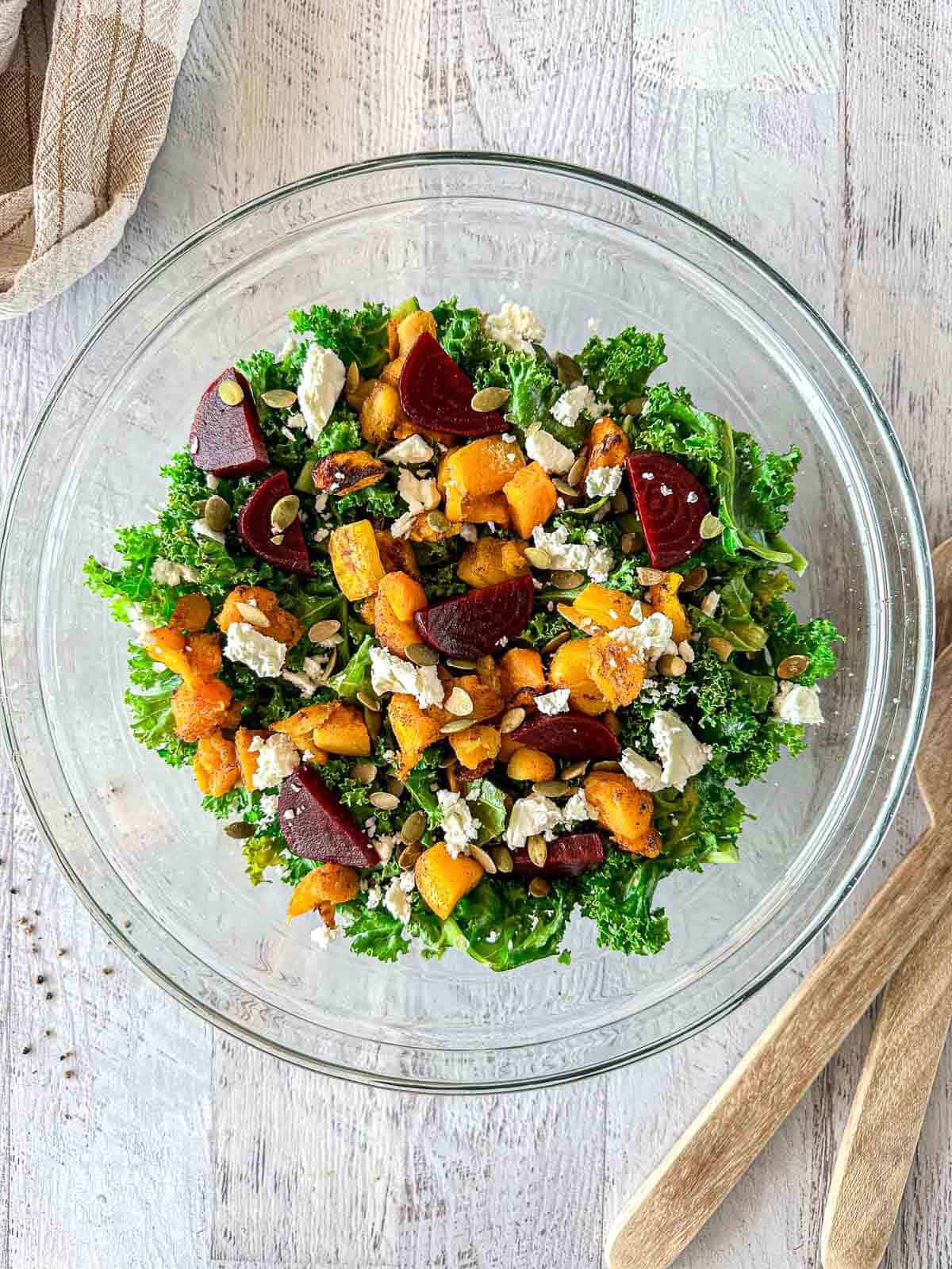 A glass bowl filled with kale salad topped with roasted butternut squash, beetroot, crumbled cheese, and pumpkin seeds sits on a light wooden surface next to wooden salad utensils.