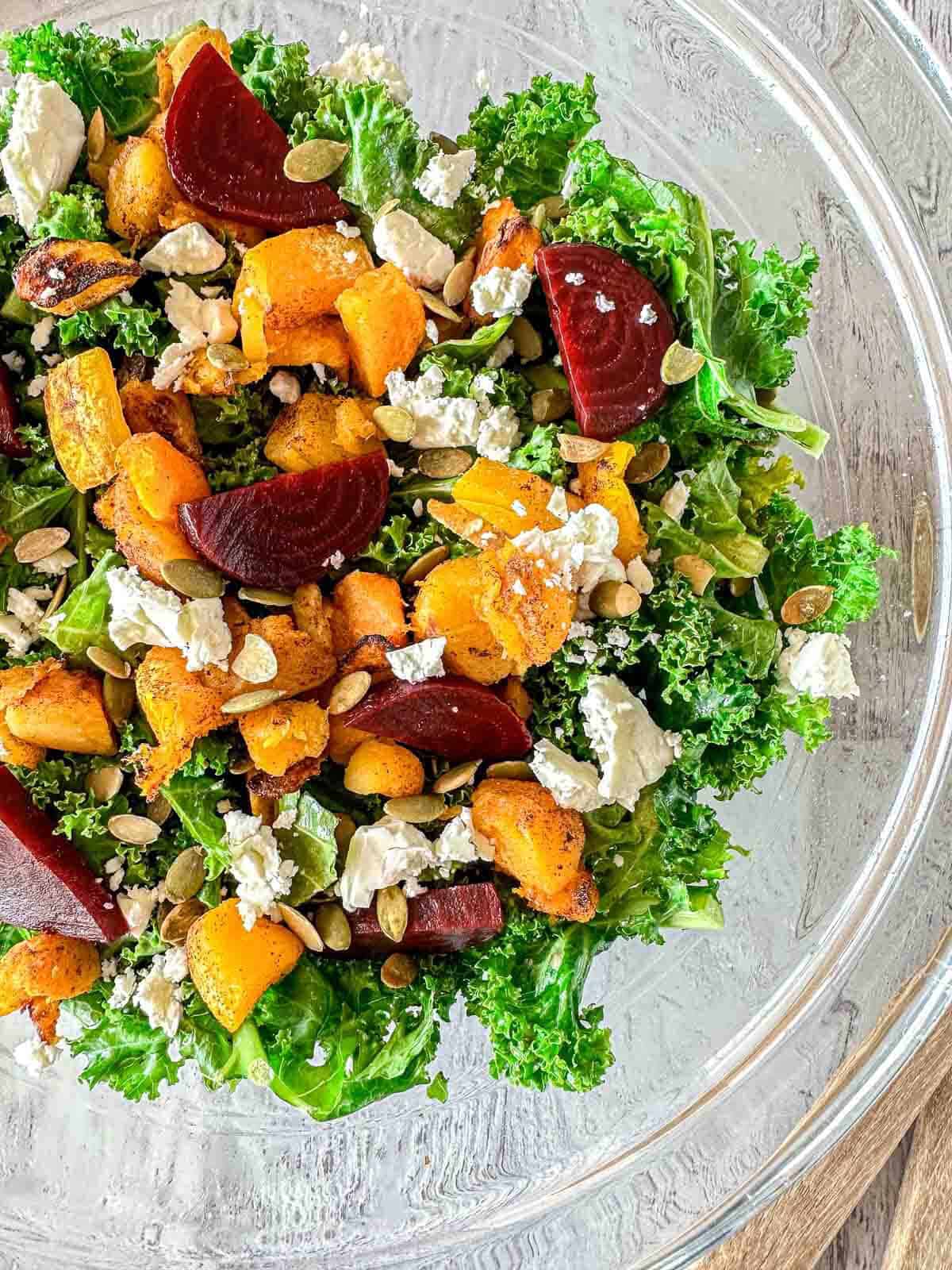 A glass bowl filled with kale salad topped with roasted butternut squash, sliced beets, crumbled feta cheese, and pumpkin seeds on a wooden surface.