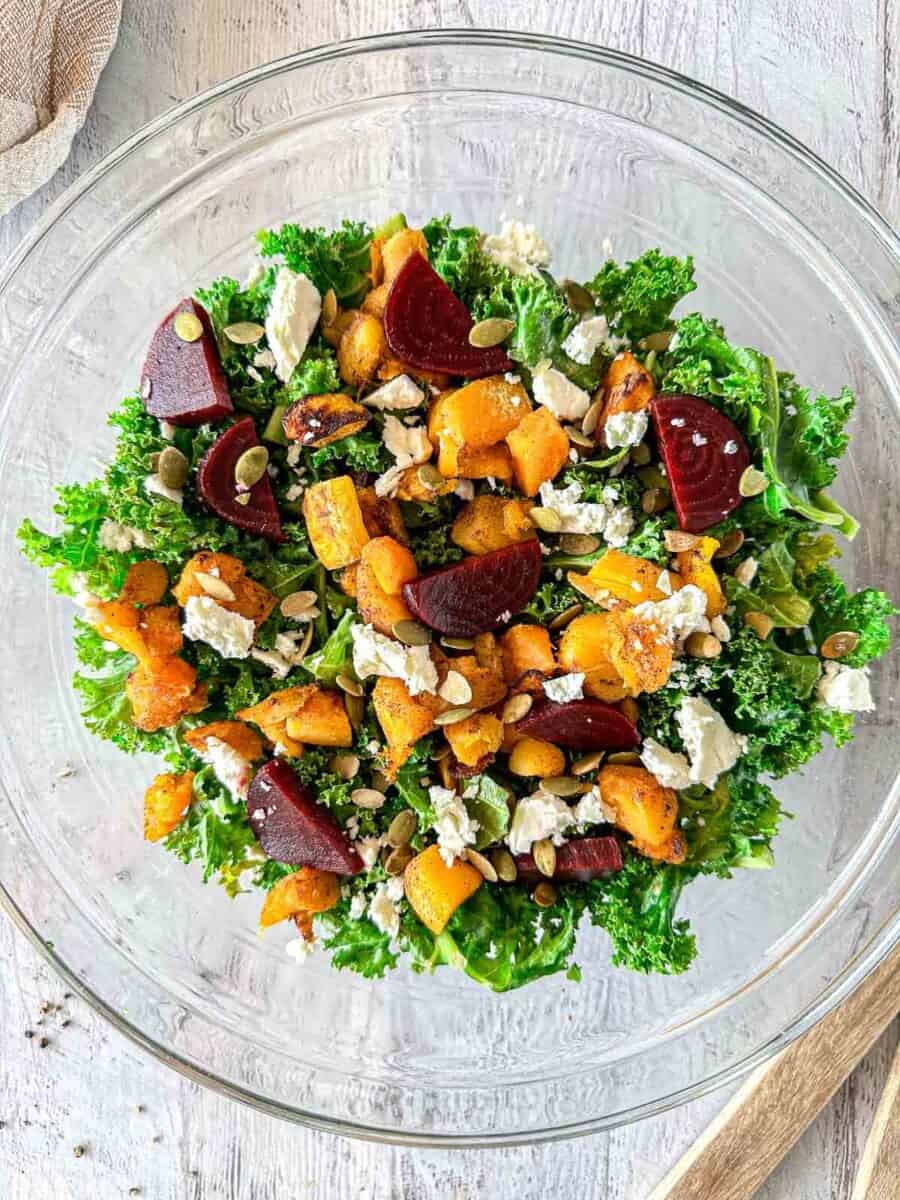 A glass bowl filled with a salad of kale, roasted butternut squash, beetroot slices, crumbled cheese, and pumpkin seeds on a light wooden surface.