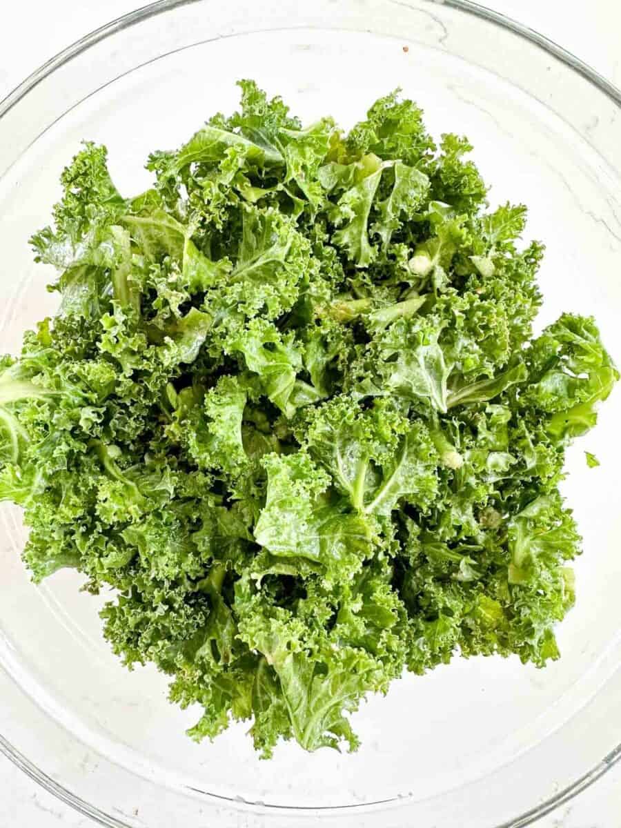 A glass bowl filled with fresh, curly green kale leaves on a white background.