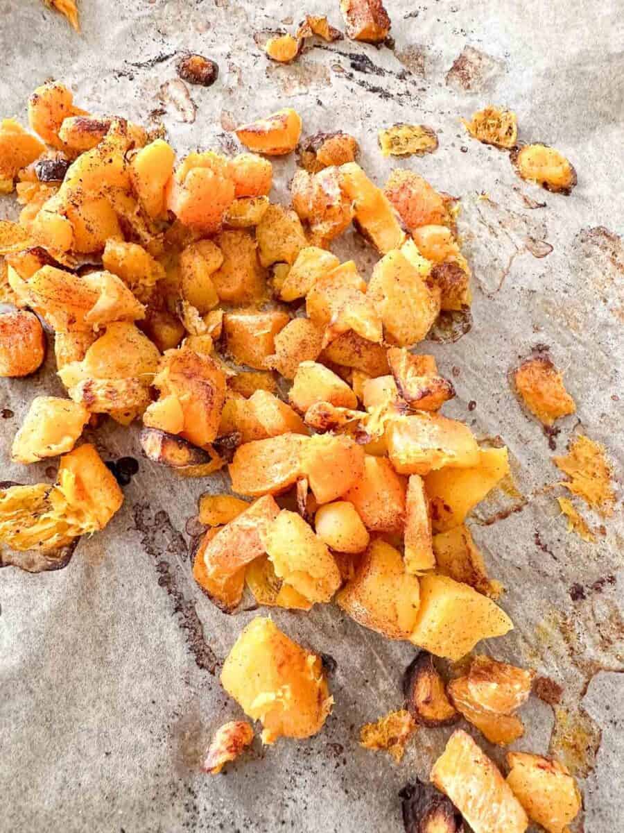 Golden, crispy roasted butternut squash scattered on a sheet of parchment paper, lightly seasoned with spices and some browned edges visible.