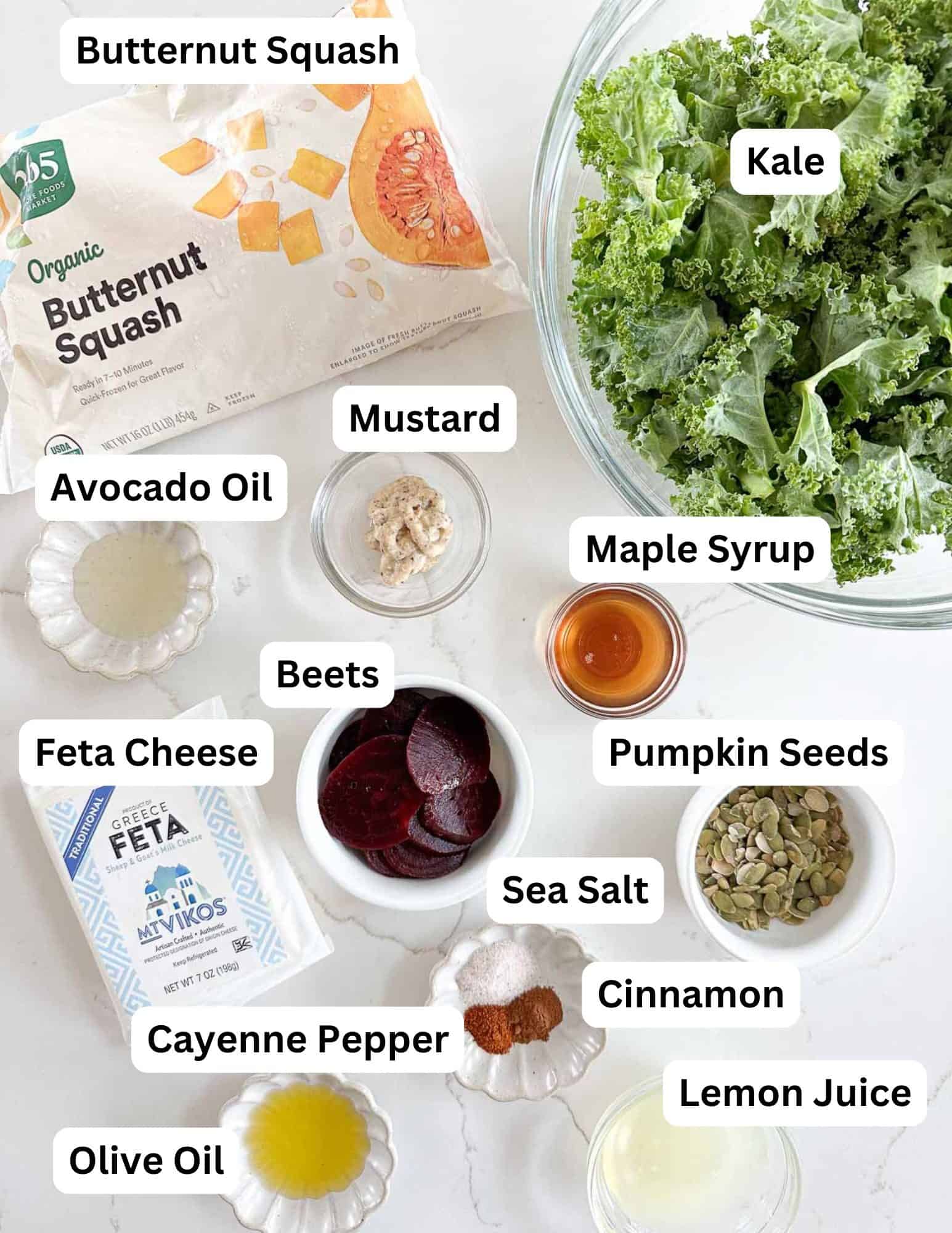 A flat lay of salad ingredients on a white surface, including labeled bowls of kale, butternut squash, avocado oil, feta cheese, beets, mustard, maple syrup, pumpkin seeds, sea salt, cinnamon, cayenne pepper, lemon juice, and olive oil.