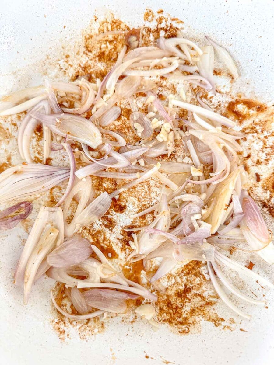 Thinly sliced shallots and minced garlic sautéing in a white pan, with brown spices scattered around, beginning to cook.