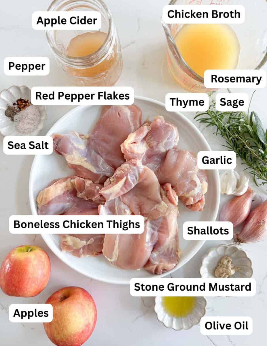 A plate of raw boneless chicken thighs is surrounded by labeled ingredients: apple cider, chicken broth, pepper, red pepper flakes, rosemary, thyme, sage, sea salt, garlic, shallots, apples, stone ground mustard, and olive oil.
