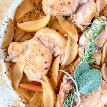 Dutch Oven Braised Chicken thighs cooked with apple slices and herbs, including a sprig of rosemary and sage, in a light sauce. The dish is served in a white baking dish, highlighting the golden chicken and tender apples.