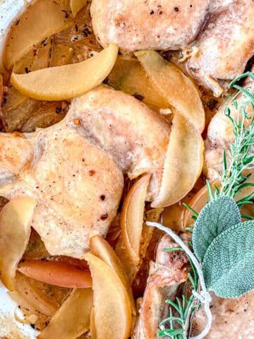 Dutch Oven Braised Chicken thighs cooked with apple slices and herbs, including a sprig of rosemary and sage, in a light sauce. The dish is served in a white baking dish, highlighting the golden chicken and tender apples.
