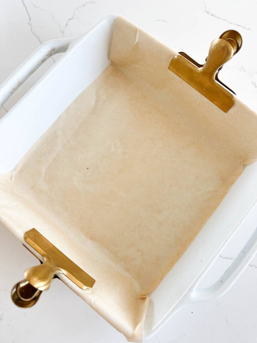 A white square baking dish lined with parchment paper, held in place at two corners by large gold binder clips, sits on a white countertop.