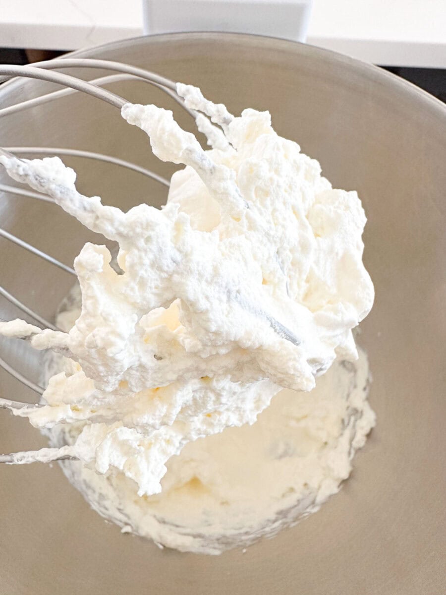 A metal whisk covered in freshly whipped cream is held over a mixing bowl containing more whipped cream.