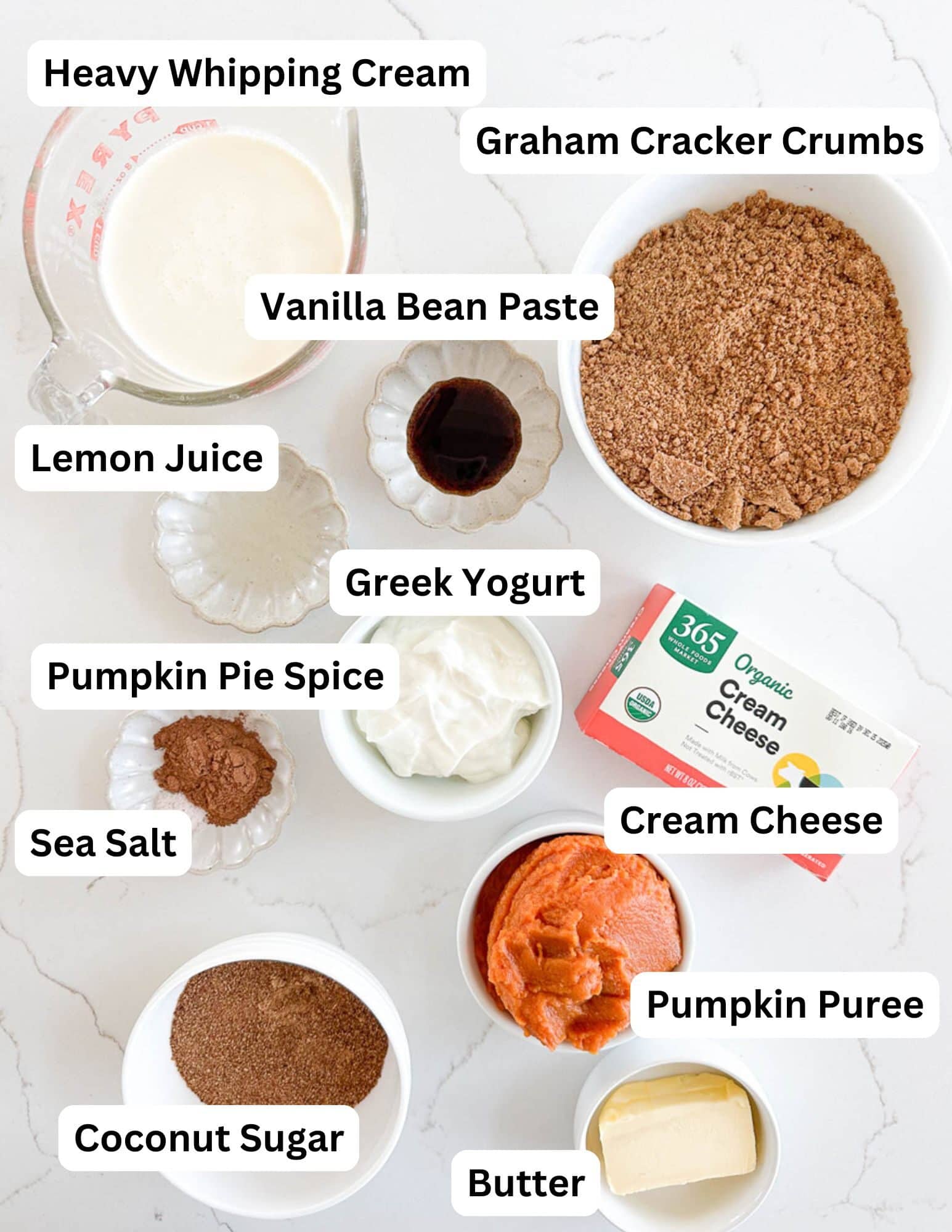 Ingredients for a pumpkin cheesecake recipe are arranged on a white surface, each labeled: heavy whipping cream, graham cracker crumbs, vanilla bean paste, lemon juice, Greek yogurt, pumpkin pie spice, sea salt, cream cheese, pumpkin puree, coconut sugar, and butter.