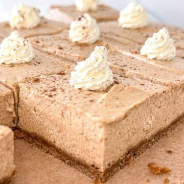 Slices of no-bake pumpkin cheesecake bars with a light brown color, each topped with a swirl of whipped cream and a sprinkle of cinnamon, on a brown parchment-lined surface.