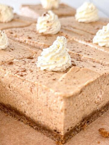 Slices of no-bake pumpkin cheesecake bars with a light brown color, each topped with a swirl of whipped cream and a sprinkle of cinnamon, on a brown parchment-lined surface.