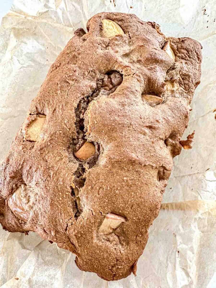 A loaf of homemade apple bread with visible apple chunks, resting on crumpled parchment paper. The bread has a golden-brown crust and a slightly cracked top.