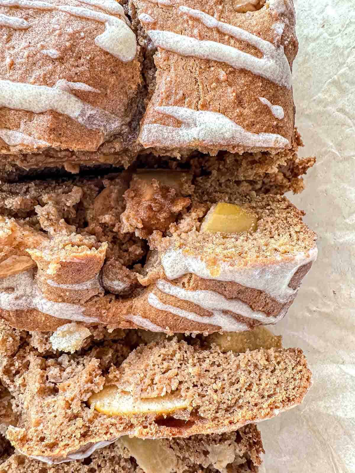 A close-up of sliced apple cinnamon bread with a light icing drizzle, showing chunks of apple and a soft, moist texture on a parchment-lined surface.