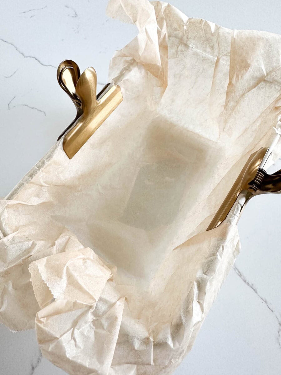 A rectangular baking dish lined with crumpled parchment paper, secured at two corners with metal clips, rests on a white marble surface.