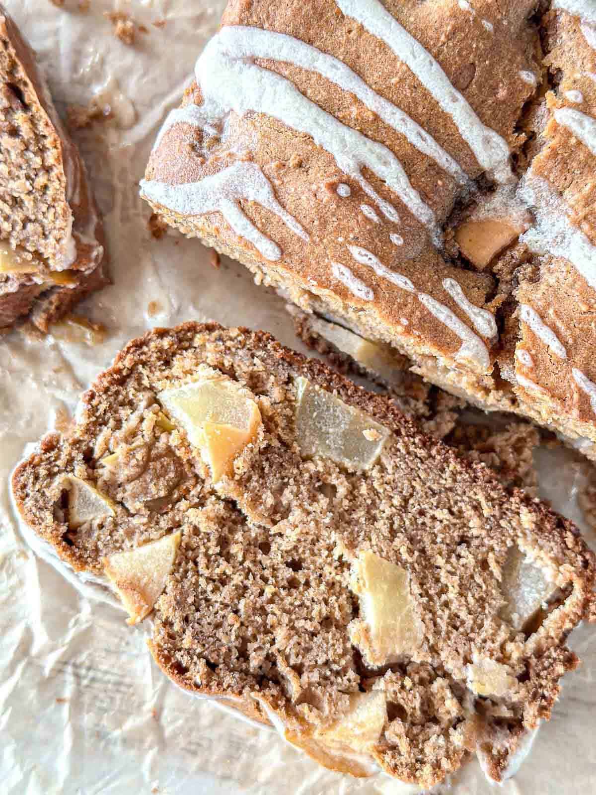 A close-up of a sliced loaf of apple cinnamon sourdough bread with visible apple chunks, drizzled with a light white glaze, resting on parchment paper.