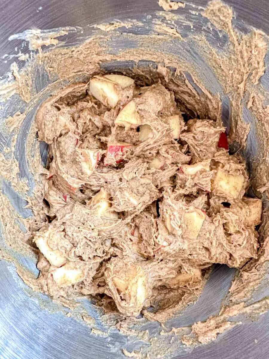 A close-up of a mixing bowl containing thick brown batter with visible chunks of chopped apples blended throughout.