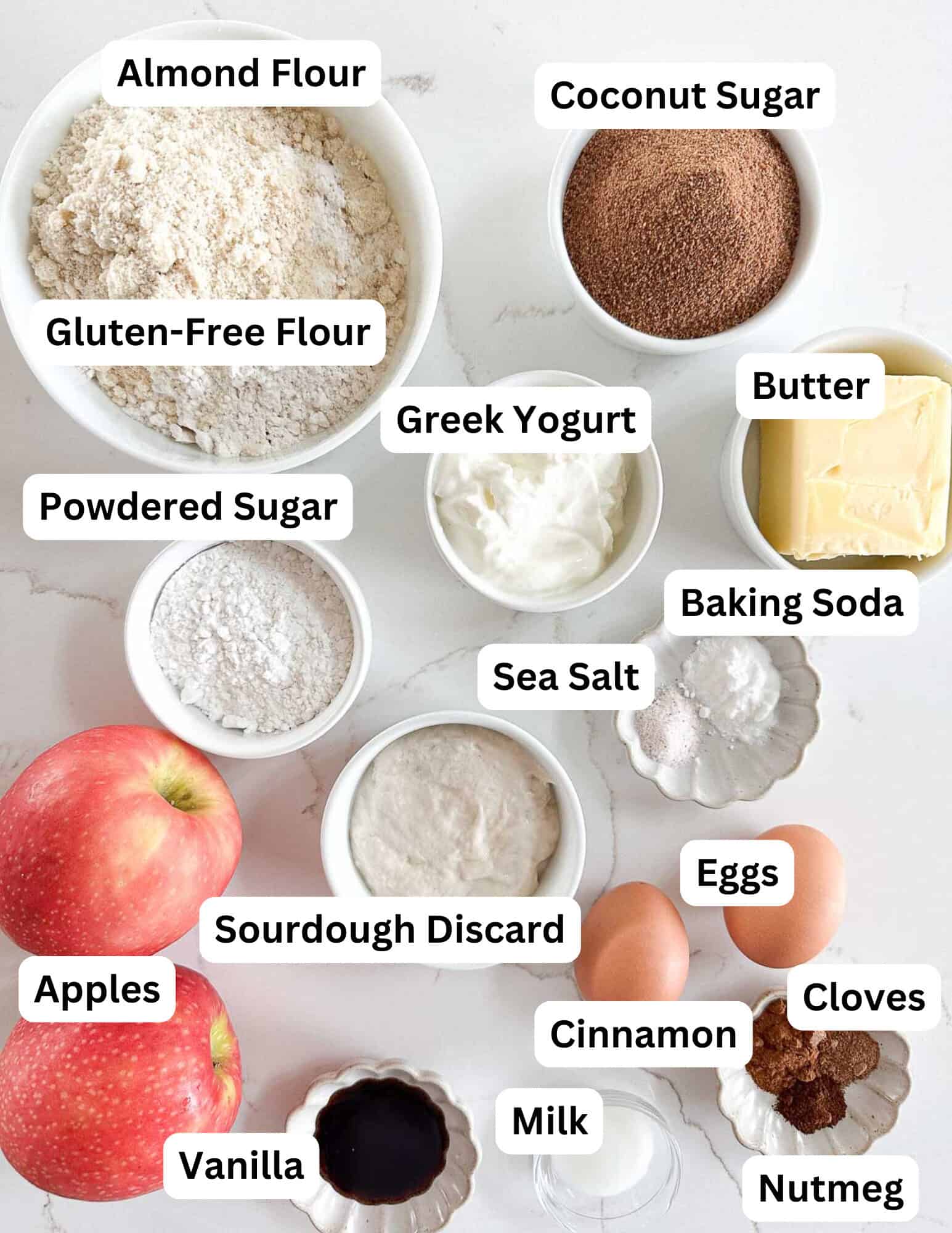A variety of labeled baking ingredients on a white surface, including bowls of almond flour, gluten-free flour, coconut sugar, powdered sugar, Greek yogurt, and butter, plus apples, eggs, and spices like cinnamon and nutmeg.