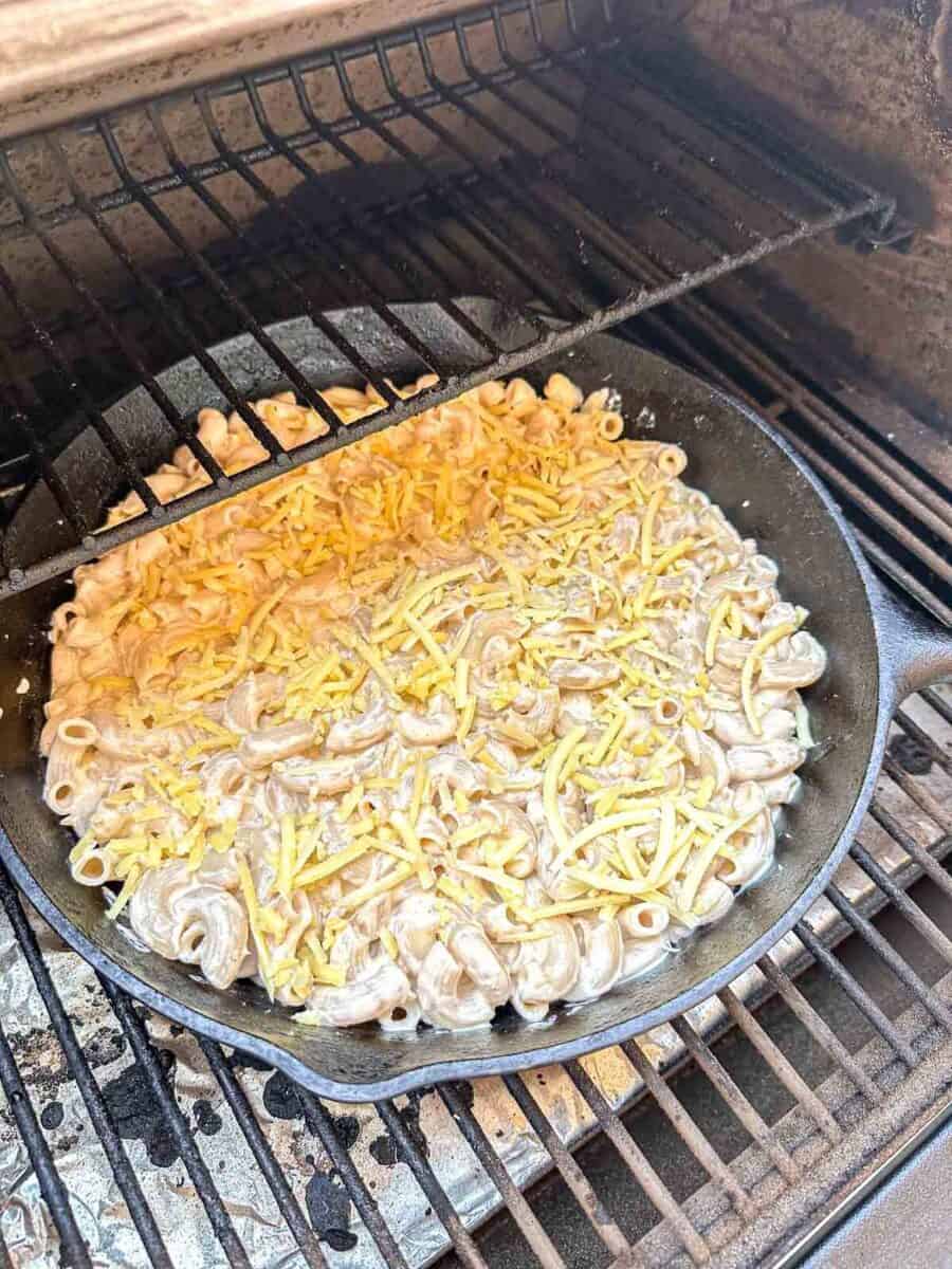 A cast iron skillet filled with macaroni and shredded cheese is placed on a grill rack, with the grill lid open, ready to be smoked.