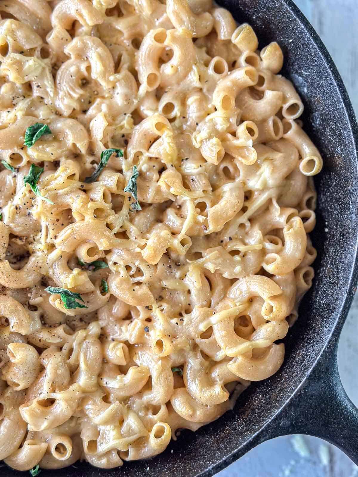 A close-up of creamy macaroni and cheese in a cast-iron skillet, topped with cracked black pepper and garnished with fresh herbs.