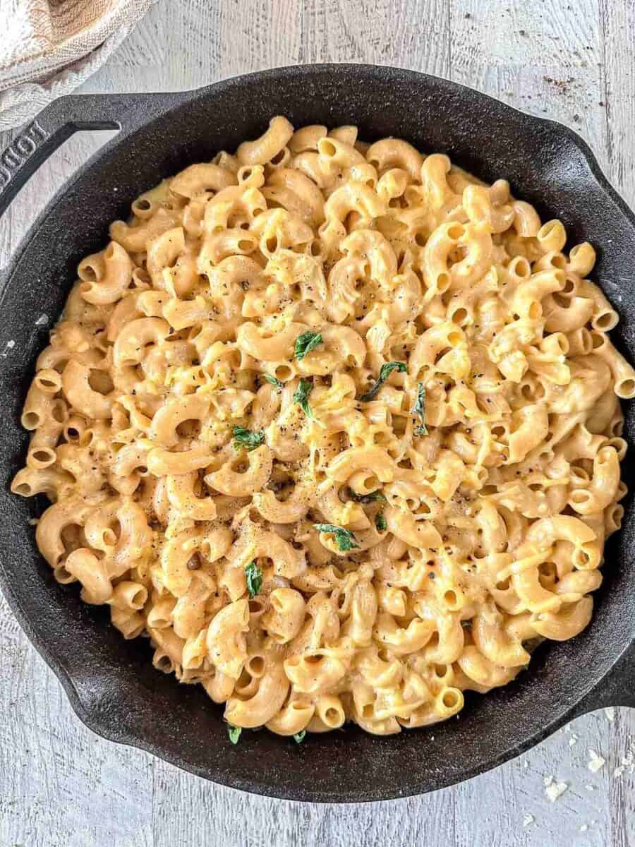 A cast iron skillet filled with creamy smoked macaroni and cheese, topped with cracked black pepper and garnished with fresh herbs, sits on a light wooden surface.
