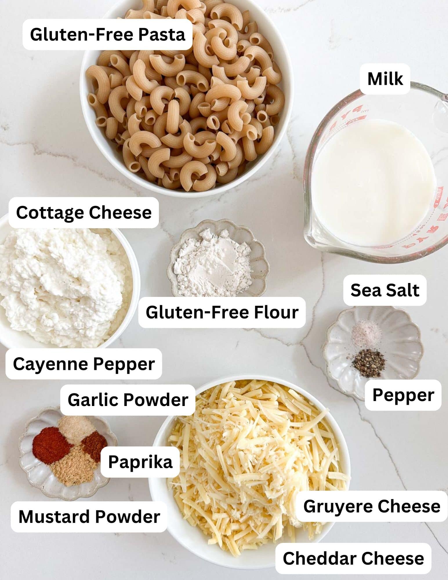Top-down view of labeled ingredients for smoked mac and cheese, including gluten-free pasta, cottage cheese, gluten-free flour, milk, sea salt, pepper, cayenne pepper, garlic powder, paprika, mustard powder, Gruyere and cheddar cheese.