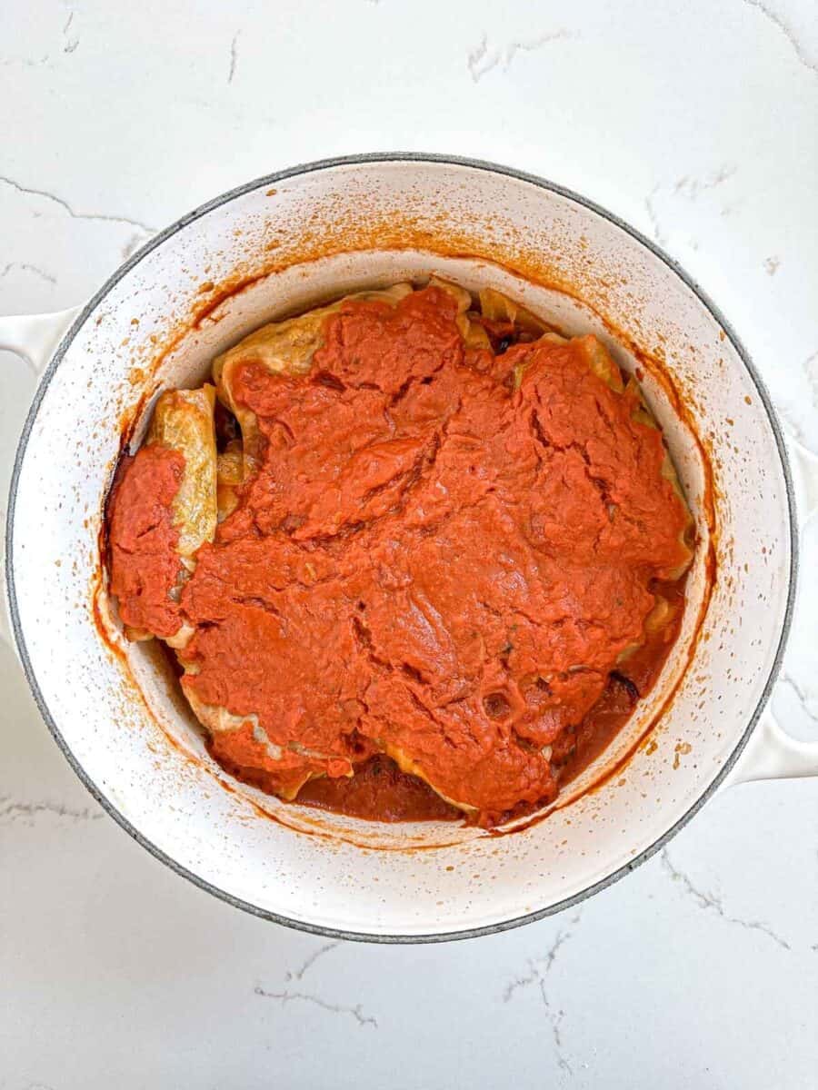 A Dutch oven filled with stuffed cabbage rolls covered in a thick red tomato sauce, sitting on a white marble surface.