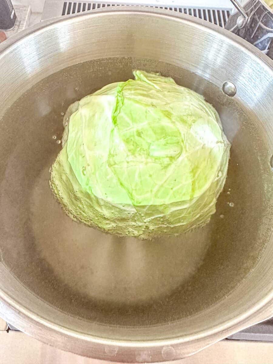 A whole head of cabbage is being boiled in a large metal pot filled with water on a stovetop.
