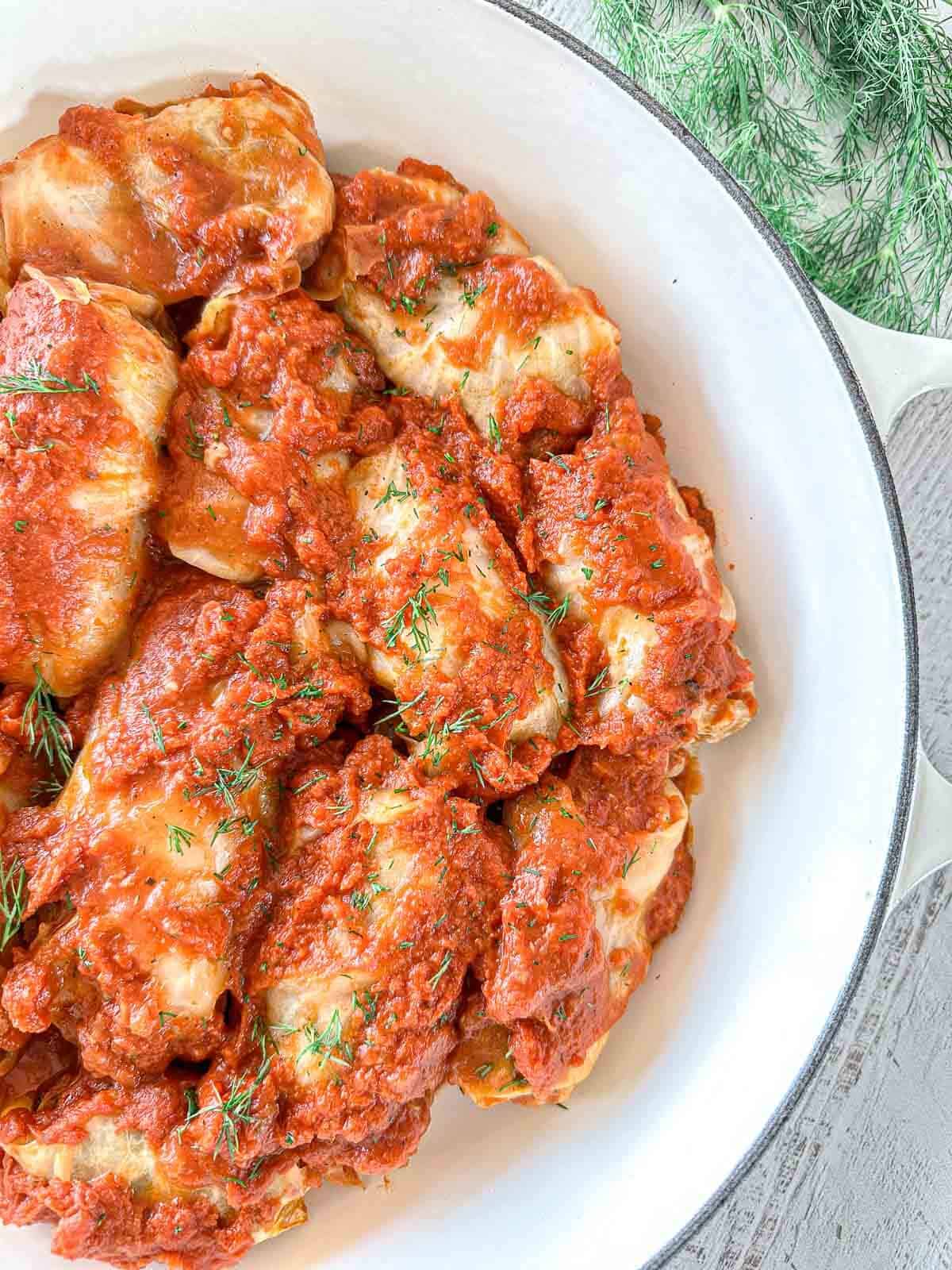 A white Dutch oven filled with cabbage rolls covered in tomato sauce and garnished with fresh dill sprigs, placed on a light-colored wooden table next to fresh dill.