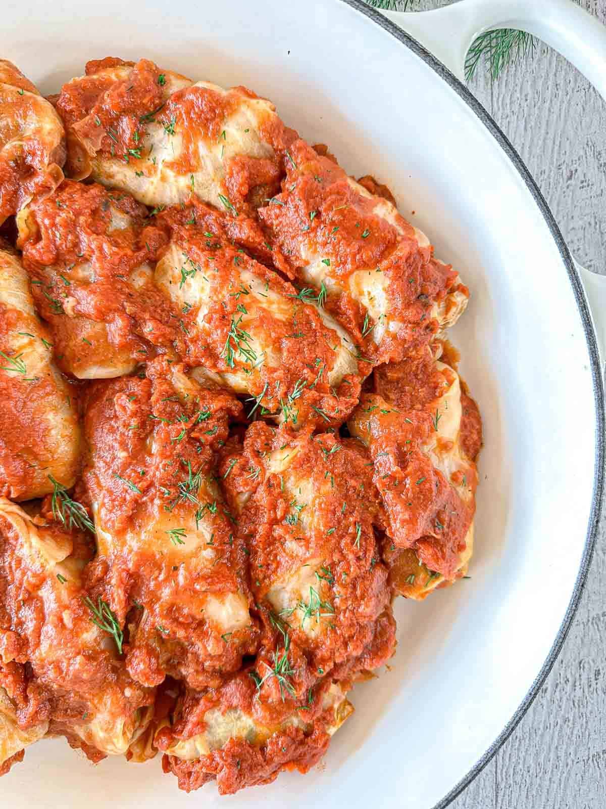 A white dish filled with cabbage rolls topped with tomato sauce and garnished with fresh herbs, placed on a light-colored surface.