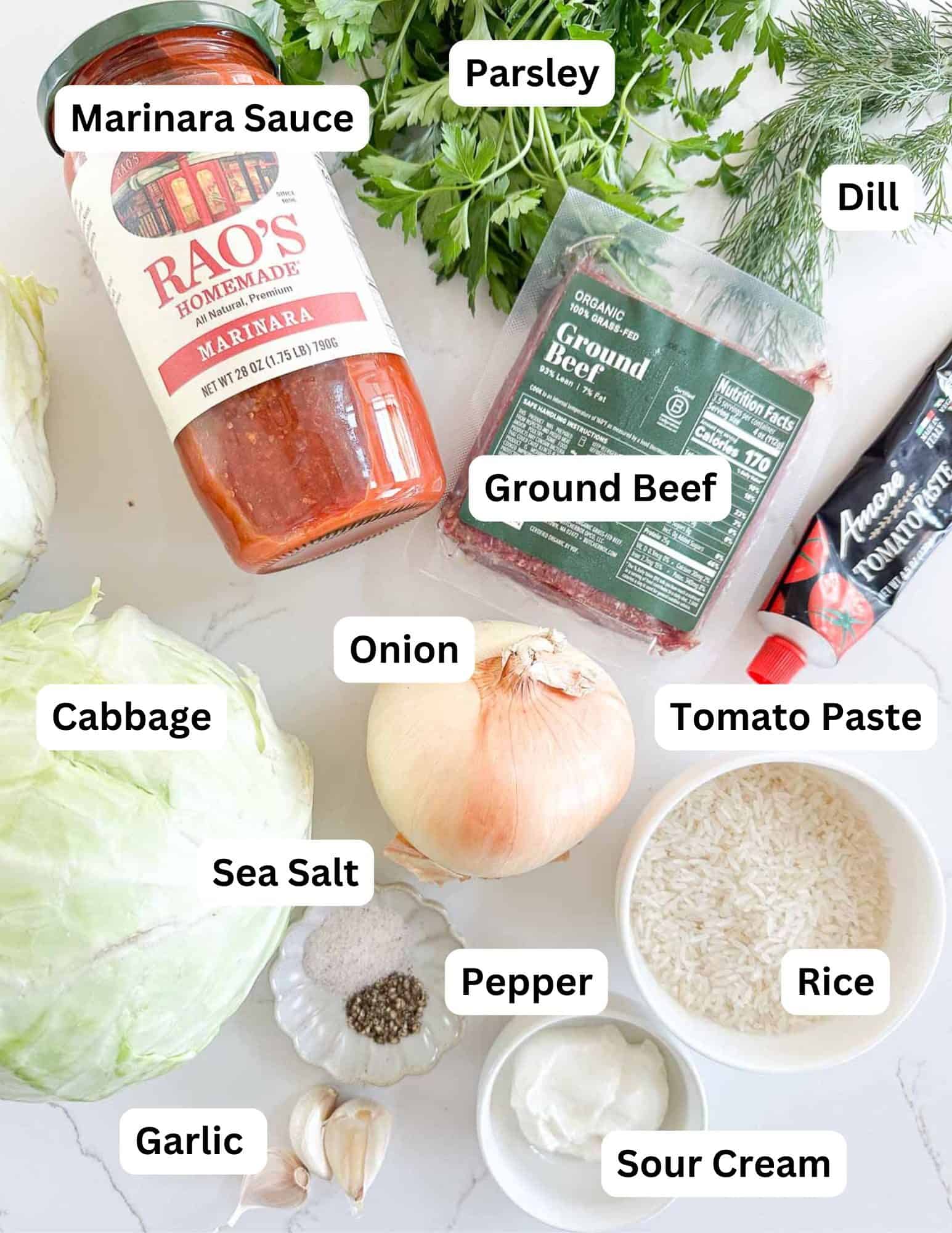 Labeled ingredients on a white surface for a recipe: marinara sauce, parsley, dill, ground beef, cabbage, onion, tomato paste, sea salt, pepper, rice, sour cream, and garlic.