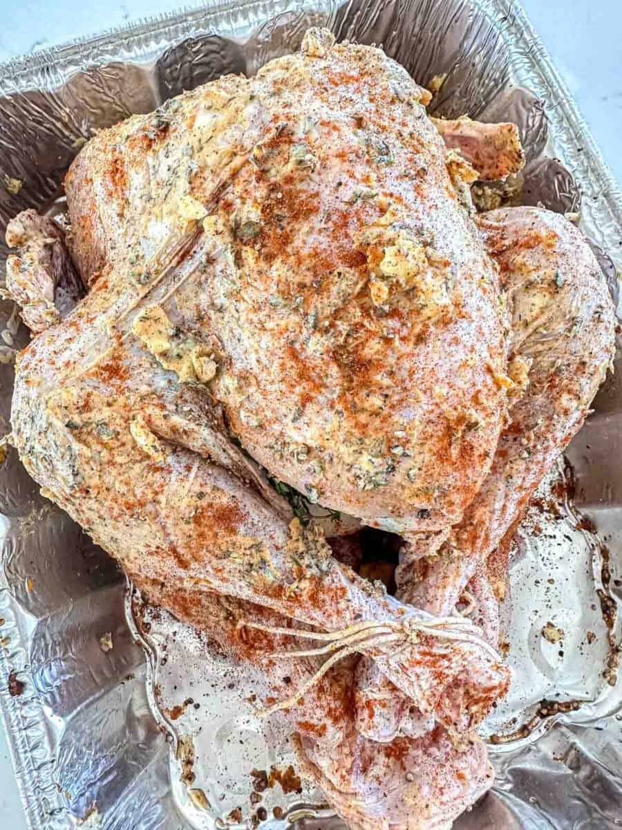 A raw whole turkey in a disposable aluminum tray, coated with a mixture of spices, herbs, and butter, ready to be cooked.