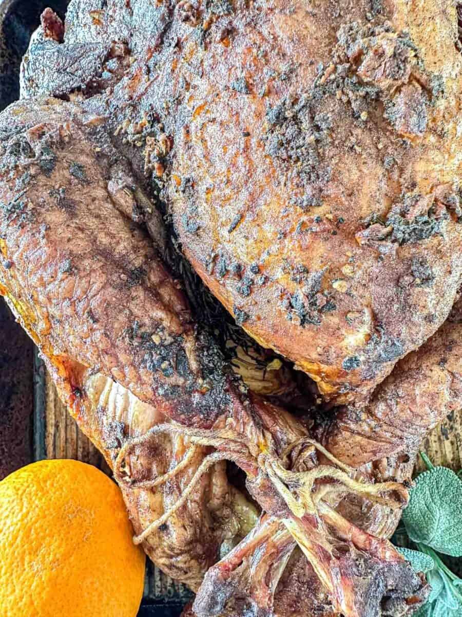 Close-up of a roasted turkey with crispy, seasoned skin, tied legs, and an orange placed nearby on a wooden surface.